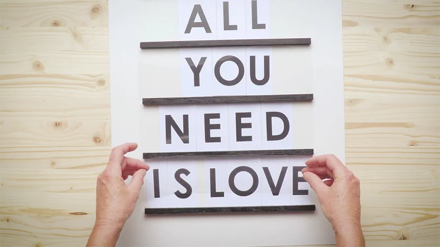 Zwei Hände befestigen schwarze Holzlatten mit dem Schriftzug 'All you need is love' auf einer weißen Fläche.