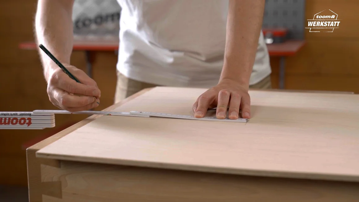 Person zeichnet mit Bleistift an einem Zollstock entlang eine Linie auf eine Holzplatte.