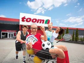 Vier lachende Personen mit Sportartikeln und Vereinsscheinen in einem Einkaufswagen.