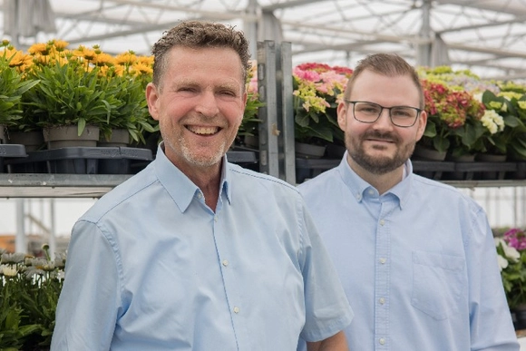 Zwei Personen stehen lächelnd vor Blumen in einem Gewächshaus.
