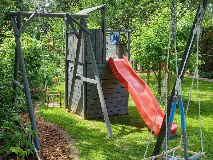 Ein graues Holzspielhaus mit Rutsche und zwei Schaukeln.