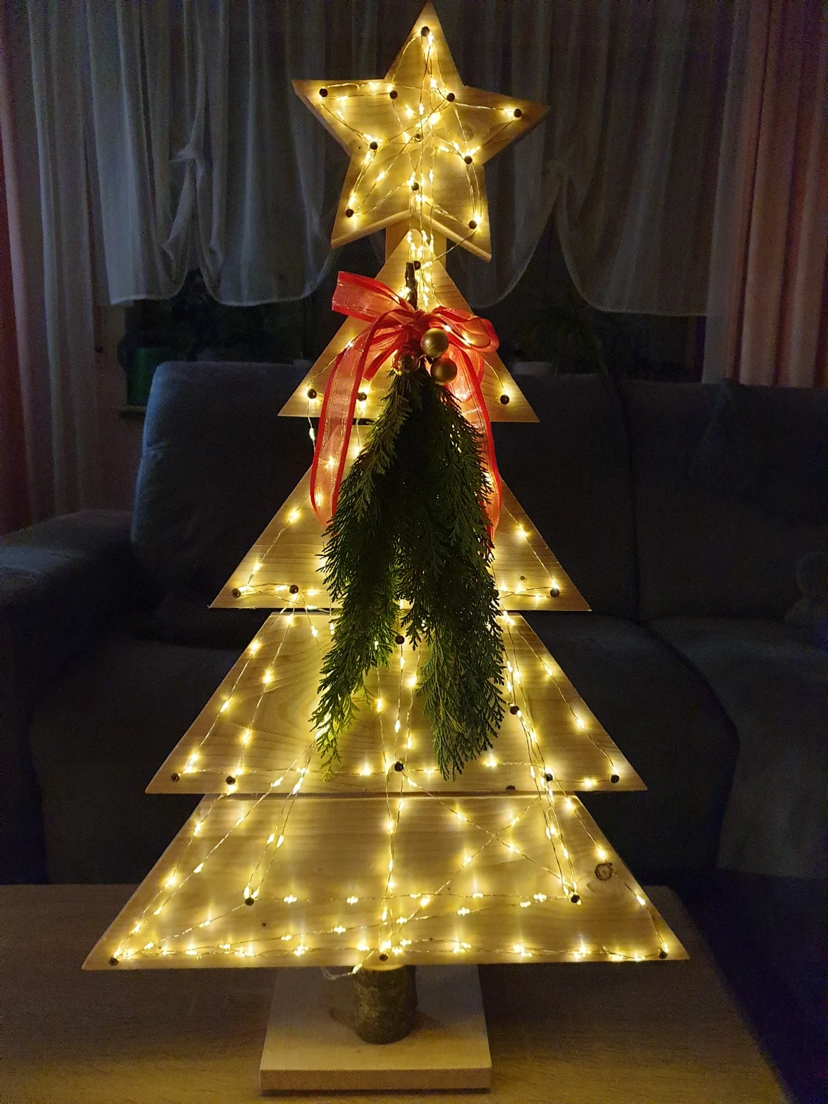 Ein beleuchteter Weihnachtsbaum aus Holz, verziert mit einem Stern und einer Schleife.