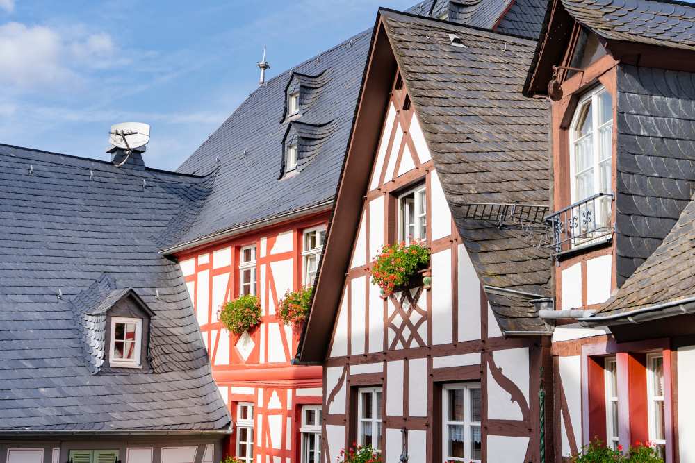 Fachwerkhäuser mit Schieferdächern und Blumenkästen vor blauem Himmel.